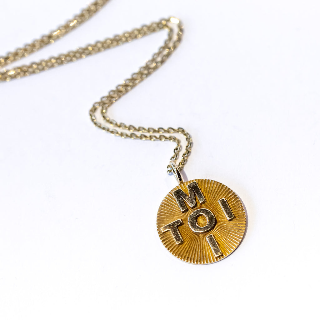 A round gold pendant with a carved starburst texture and the words "toi" and "moi" crossed in block letters through the middle. Hands from a simple gold chain necklace.
