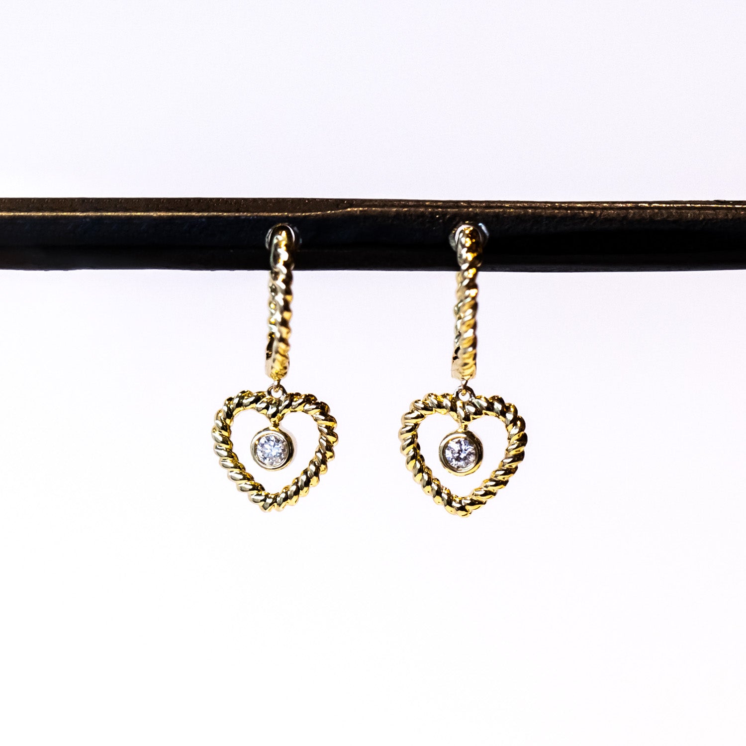 A twist-design huggie style hoop earring with a twisted wire open heart drop and a bezel set diamond dangle in the center.