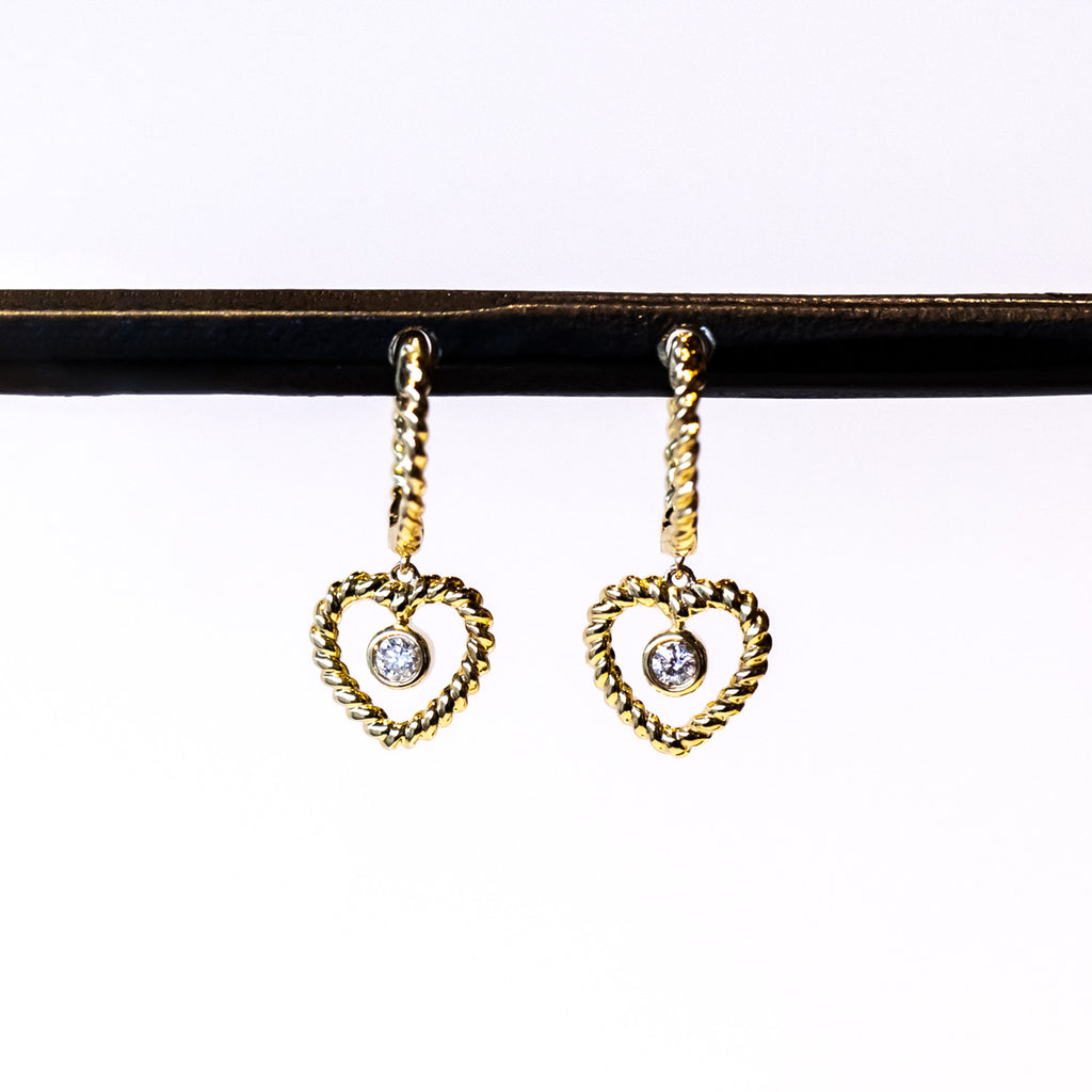 A twist-design huggie style hoop earring with a twisted wire open heart drop and a bezel set diamond dangle in the center.