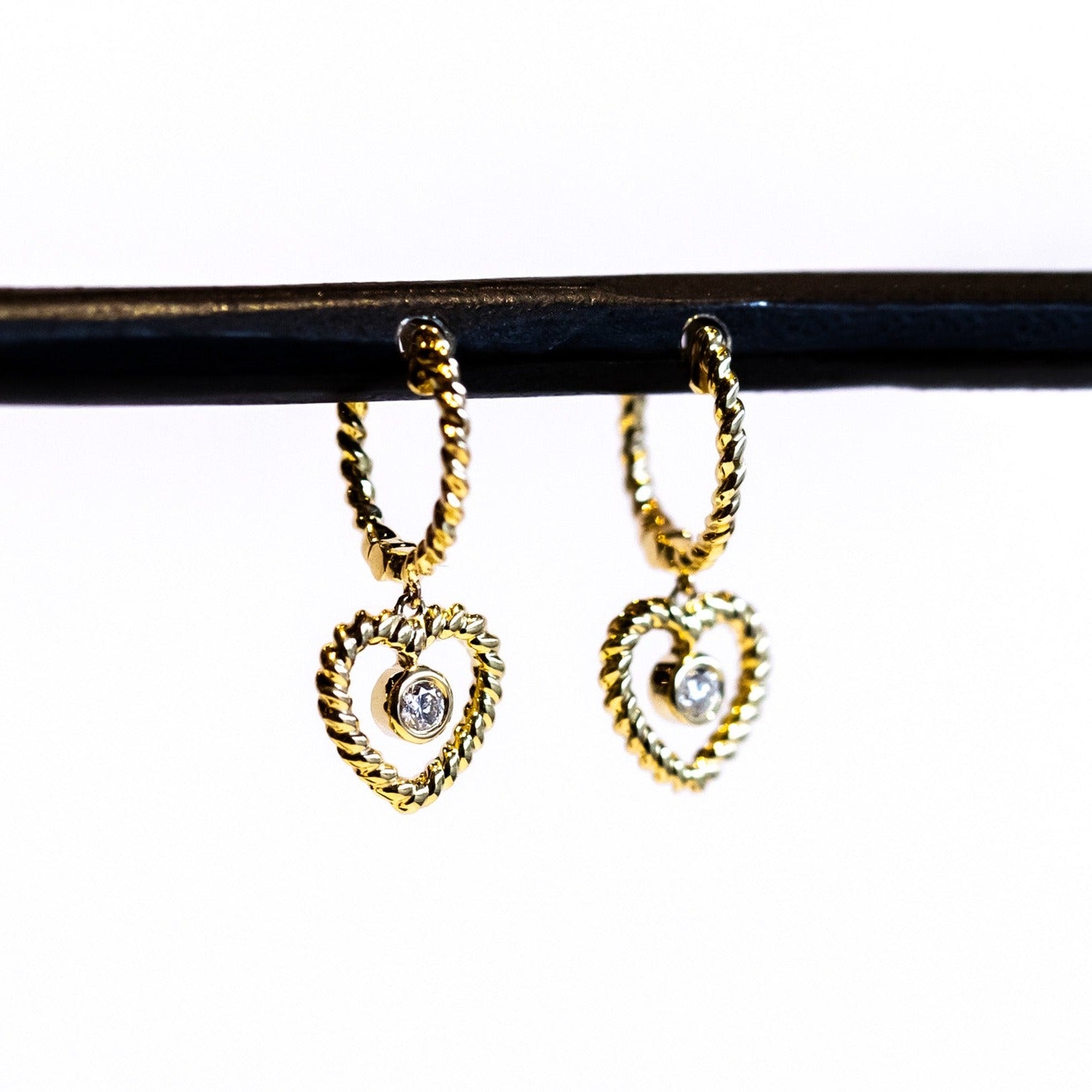 A twist-design huggie style hoop earring with a twisted wire open heart drop and a bezel set diamond dangle in the center.