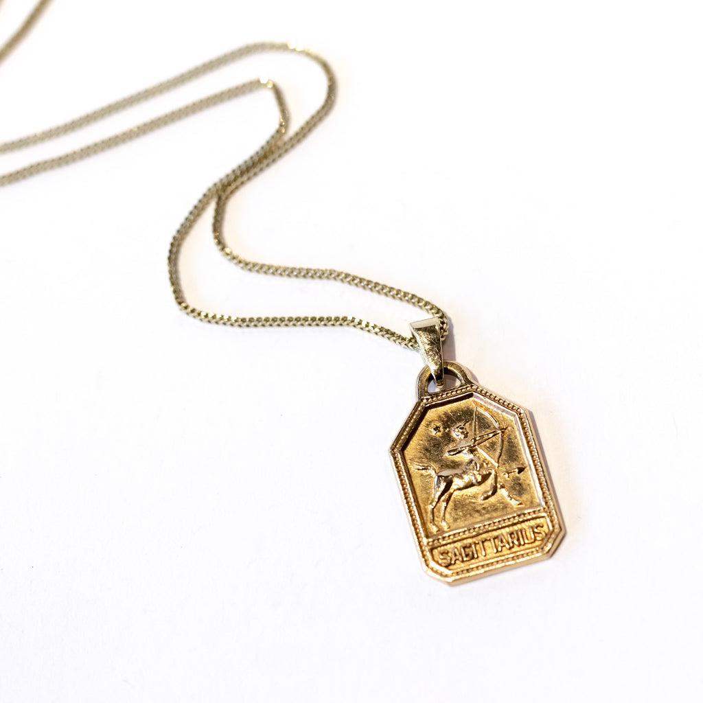 A dog tag style gold charm necklace featuring a zodiac sagittarius symbol.