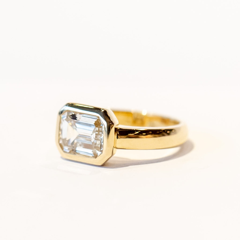 A chunky yellow gold diamond engagement ring with a bezel set emerald cut diamond set horizontally.