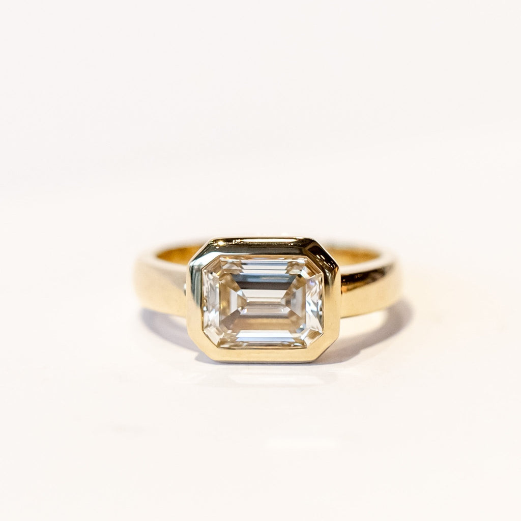 A chunky yellow gold diamond engagement ring with a bezel set emerald cut diamond set horizontally.
