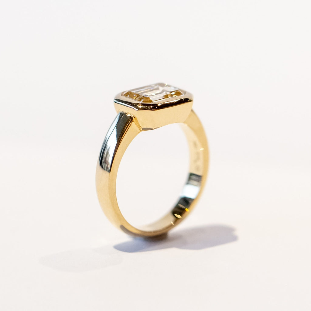 A chunky yellow gold diamond engagement ring with a bezel set emerald cut diamond set horizontally.