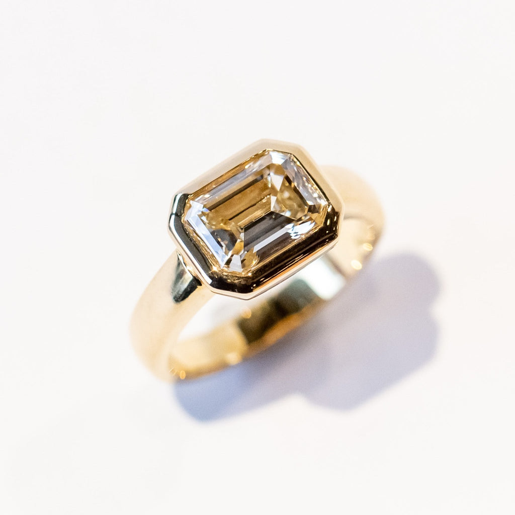 A chunky yellow gold diamond engagement ring with a bezel set emerald cut diamond set horizontally.