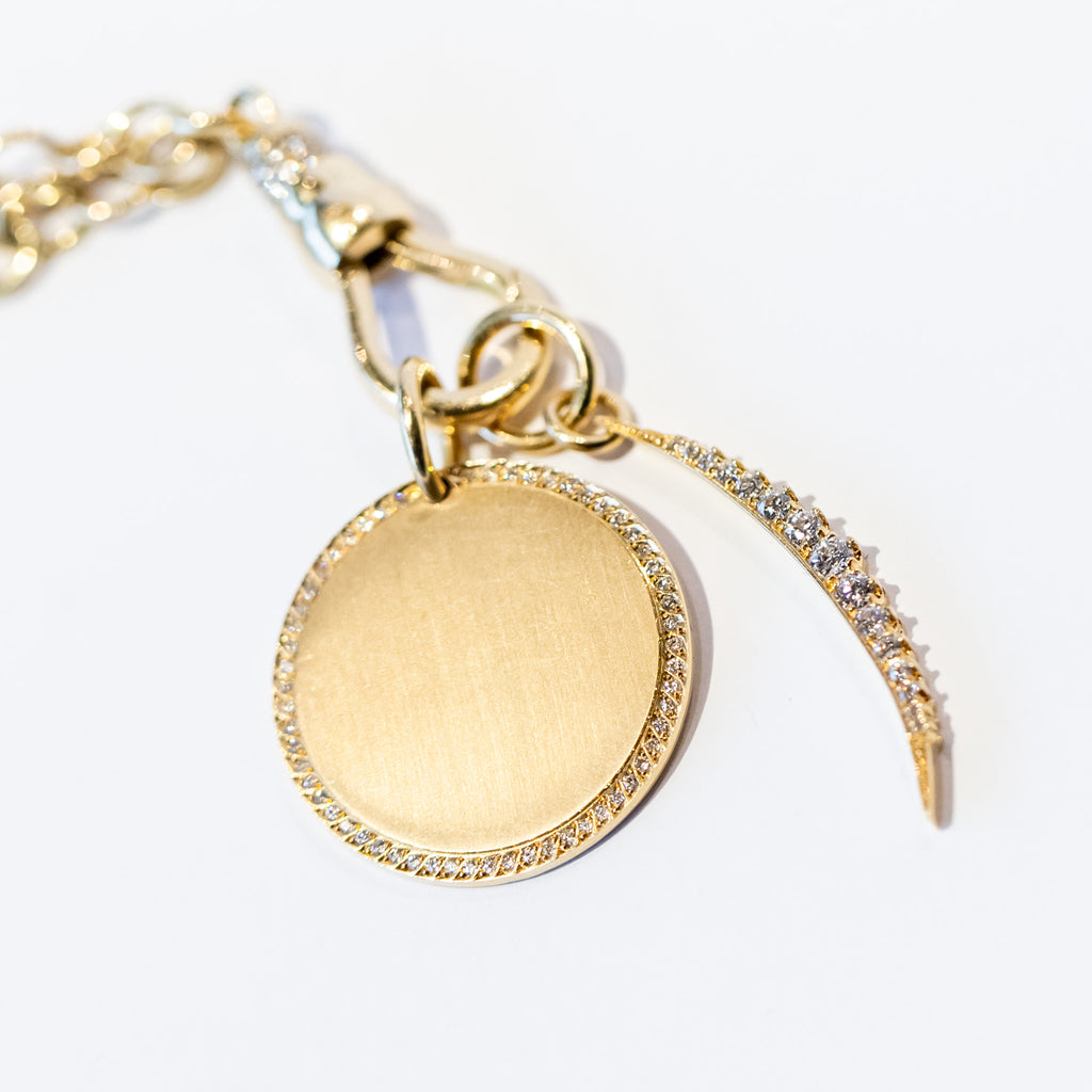 A slim, elongated crescent moon charm in yellow gold, set with a row of pave diamonds, accompanied by a large round circle charm on a gold chain.