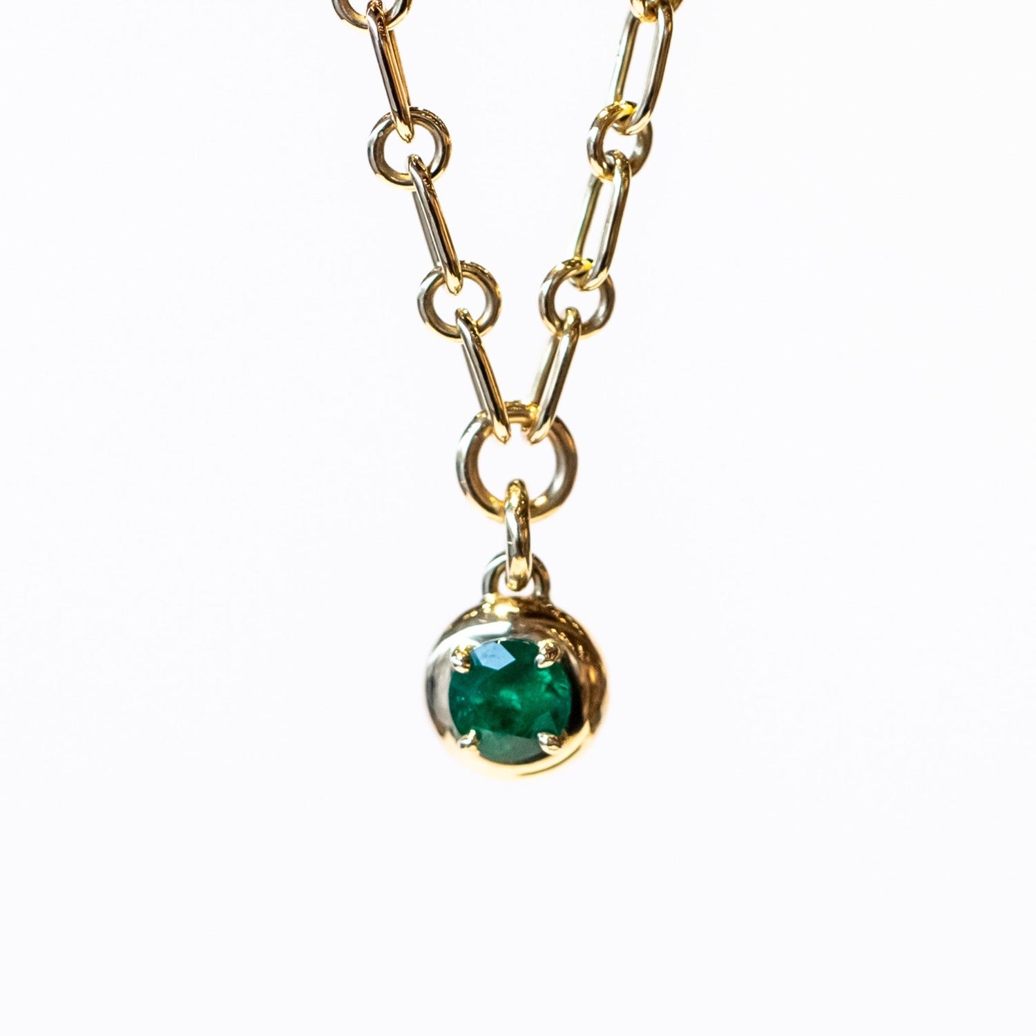 A gold chain necklace with a gold and emerald charm. The round charm features a round green emerald in a prong setting.