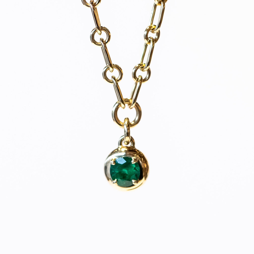 A gold chain necklace with a gold and emerald charm. The round charm features a round green emerald in a prong setting.