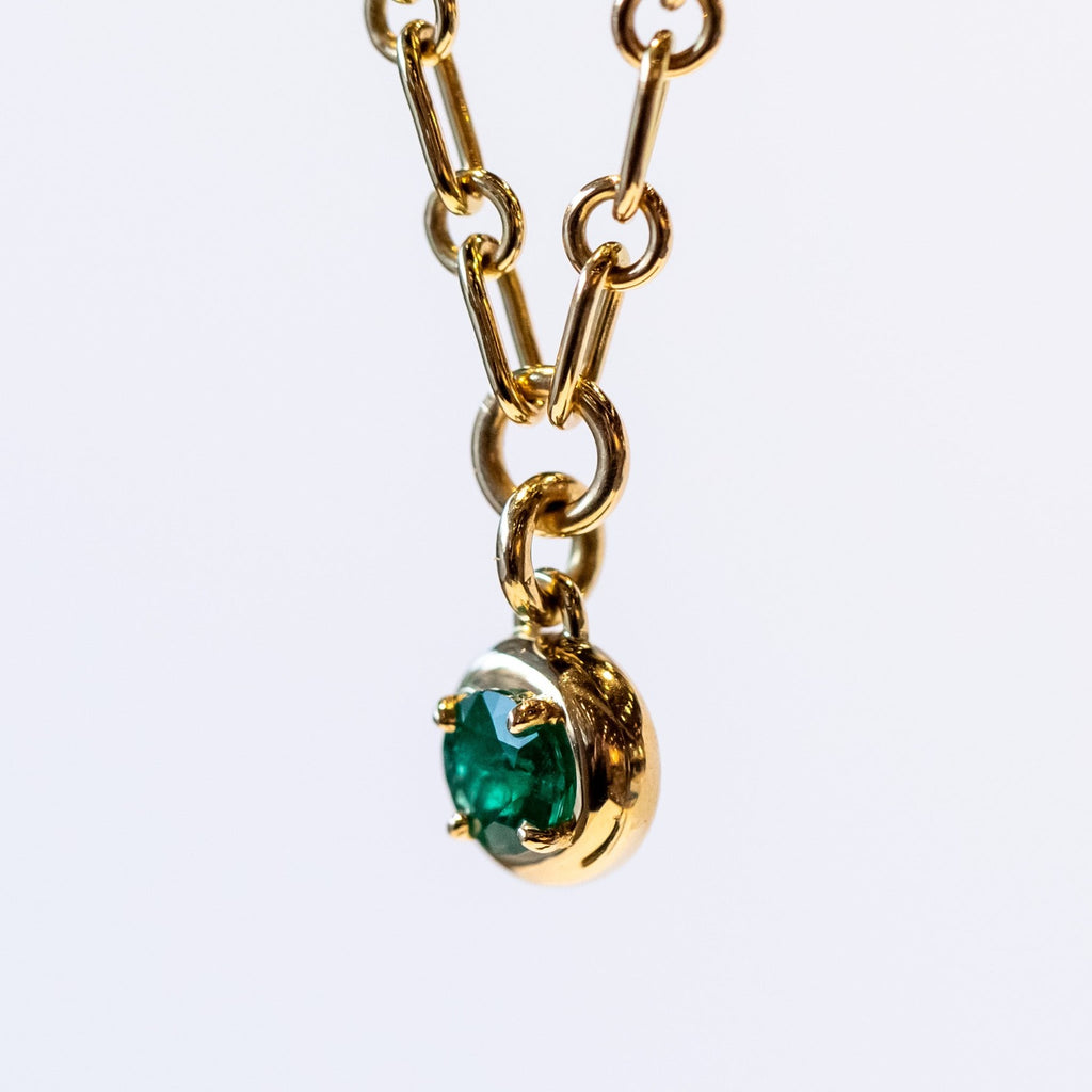 A gold chain necklace with a gold and emerald charm. The round charm features a round green emerald in a prong setting.
