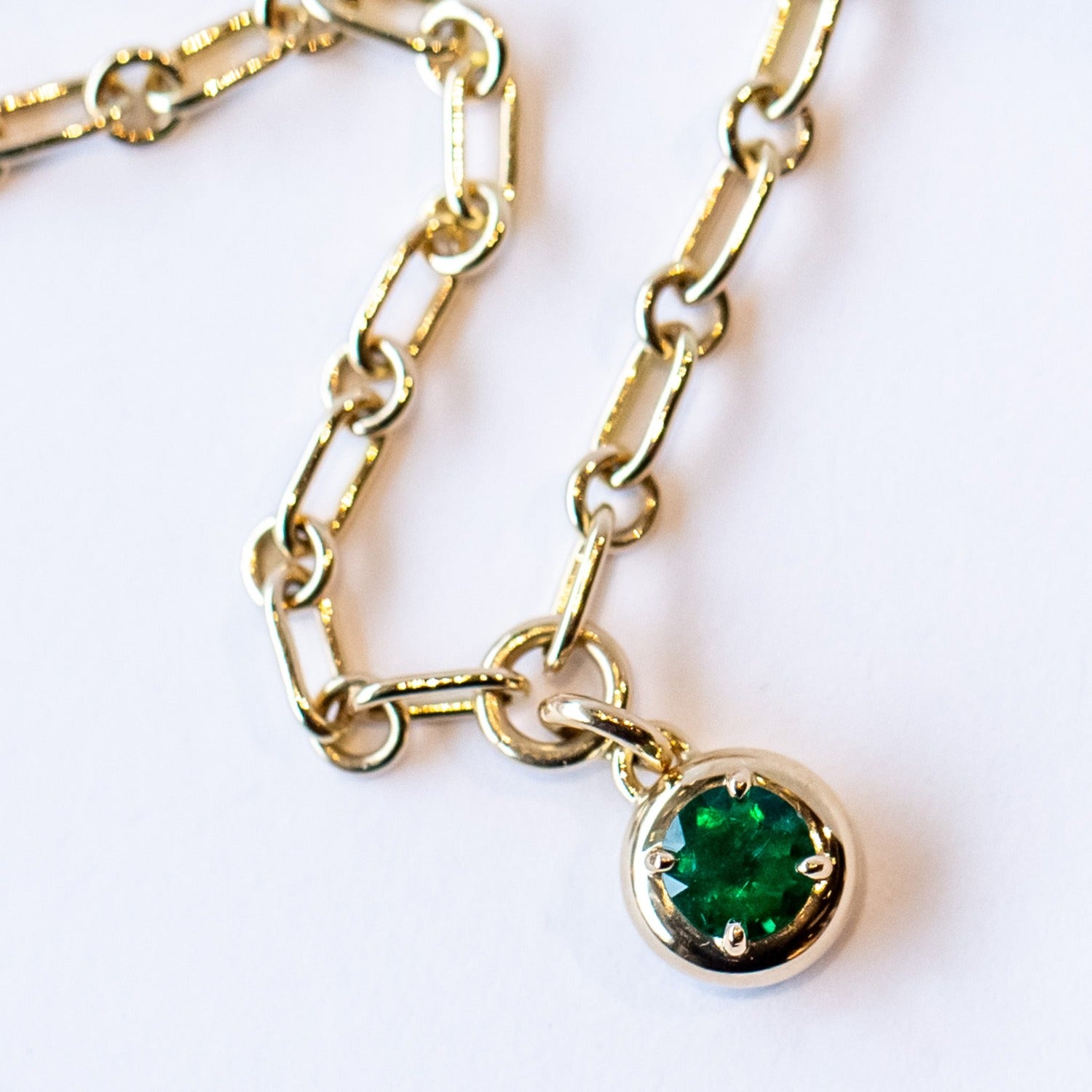 A gold chain necklace with a gold and emerald charm. The round charm features a round green emerald in a prong setting.