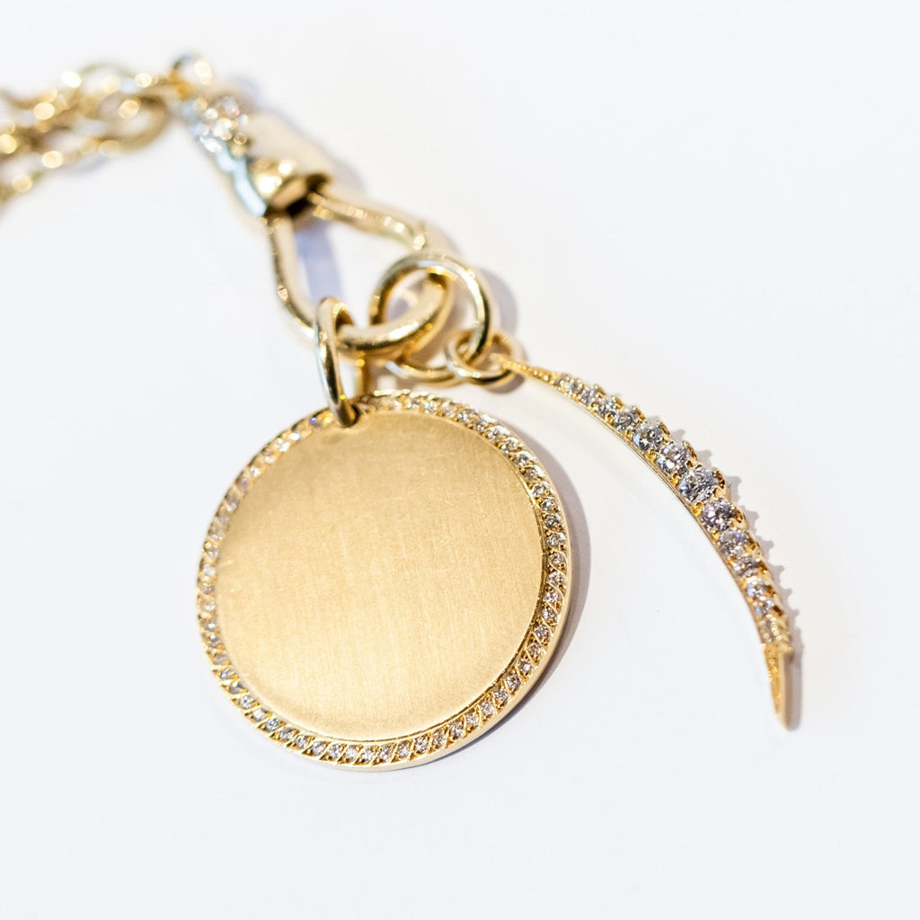 A yellow gold, round disc charm with a ring of diamonds around the outer edge and a brushed matte finish. Paired with a moon charm on a gold chain.