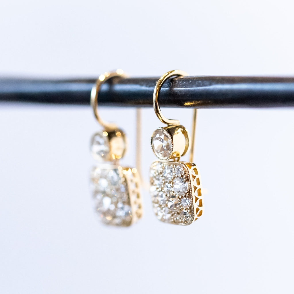 Yellow gold diamond drop earrings featuring a rounded square drop below a small circle. The top circle features one bezel set diamond while the square is covered in cobblestone pave diamonds.