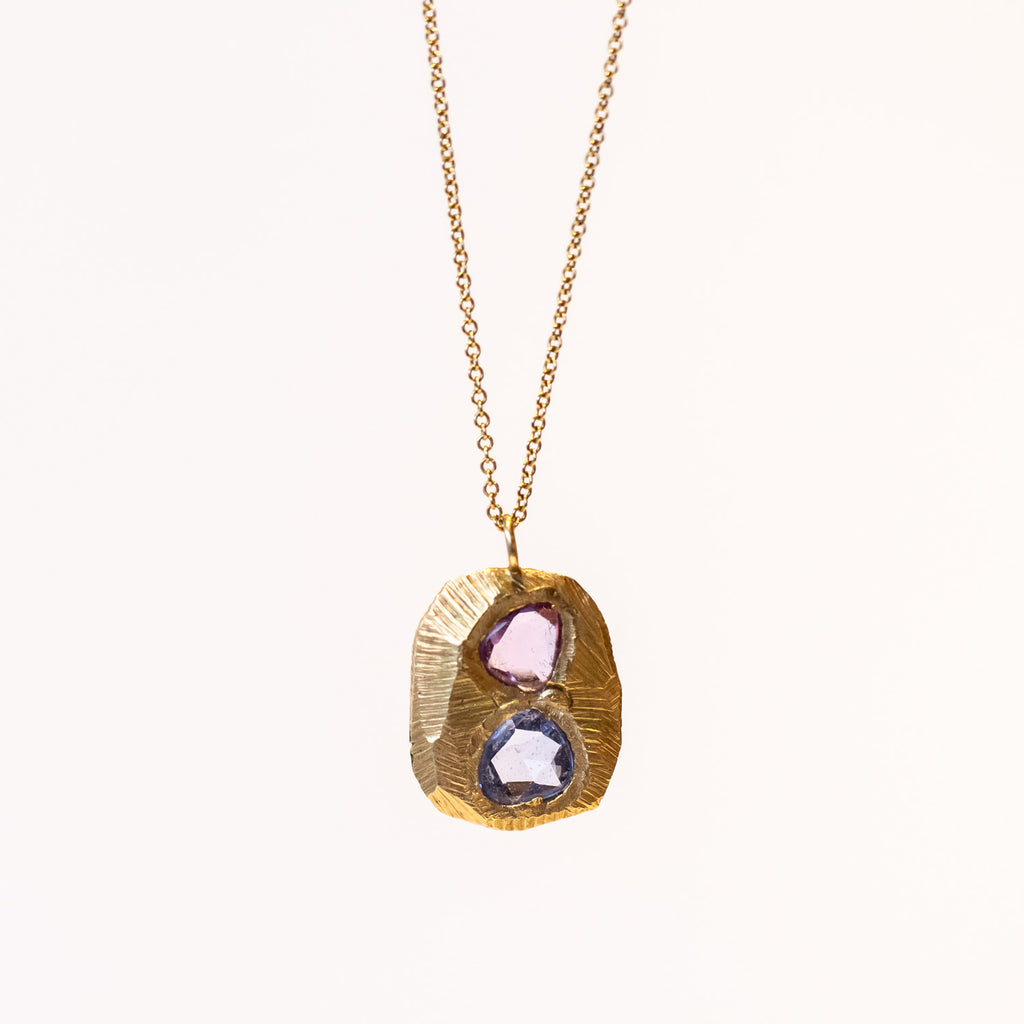 A carved yellow gold tablet is set with free form shaped sapphires, one pink, one purplish-blue on a cable chain necklace.