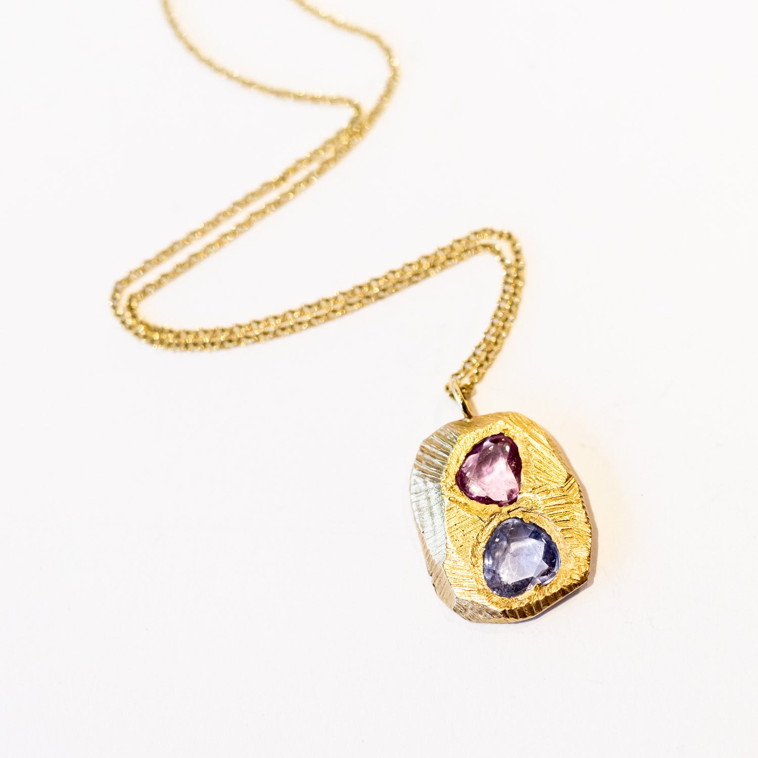 A carved yellow gold tablet is set with free form shaped sapphires, one pink, one purplish-blue on a cable chain necklace.