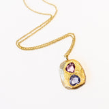 A carved yellow gold tablet is set with free form shaped sapphires, one pink, one purplish-blue on a cable chain necklace.