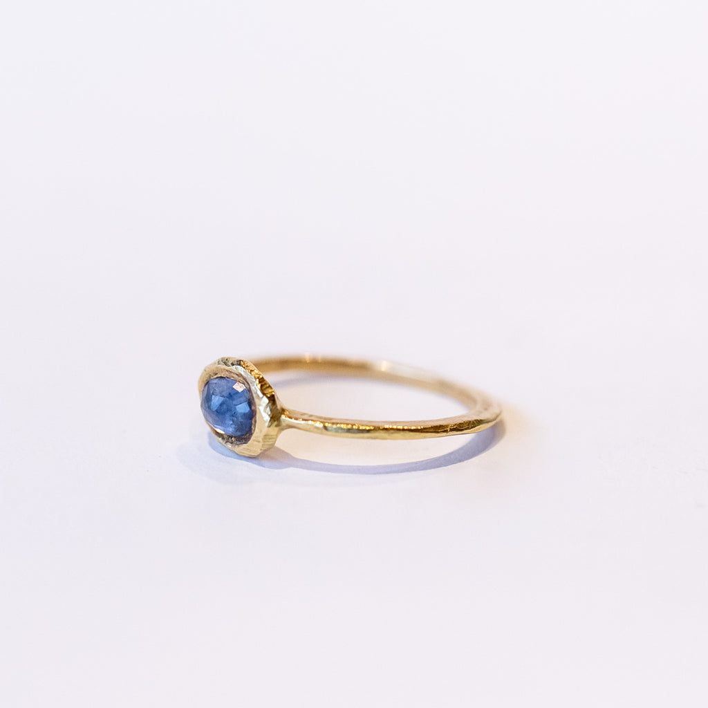 An east-west set oval rose cut blue sapphire is held in a textured yellow gold bezel on a slender ring.