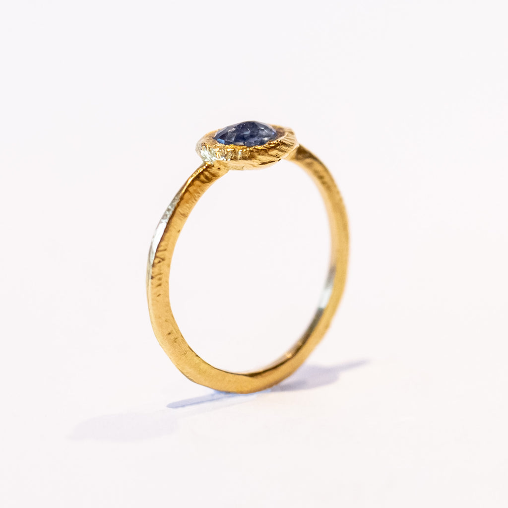 An east-west set oval rose cut blue sapphire is held in a textured yellow gold bezel on a slender ring.
