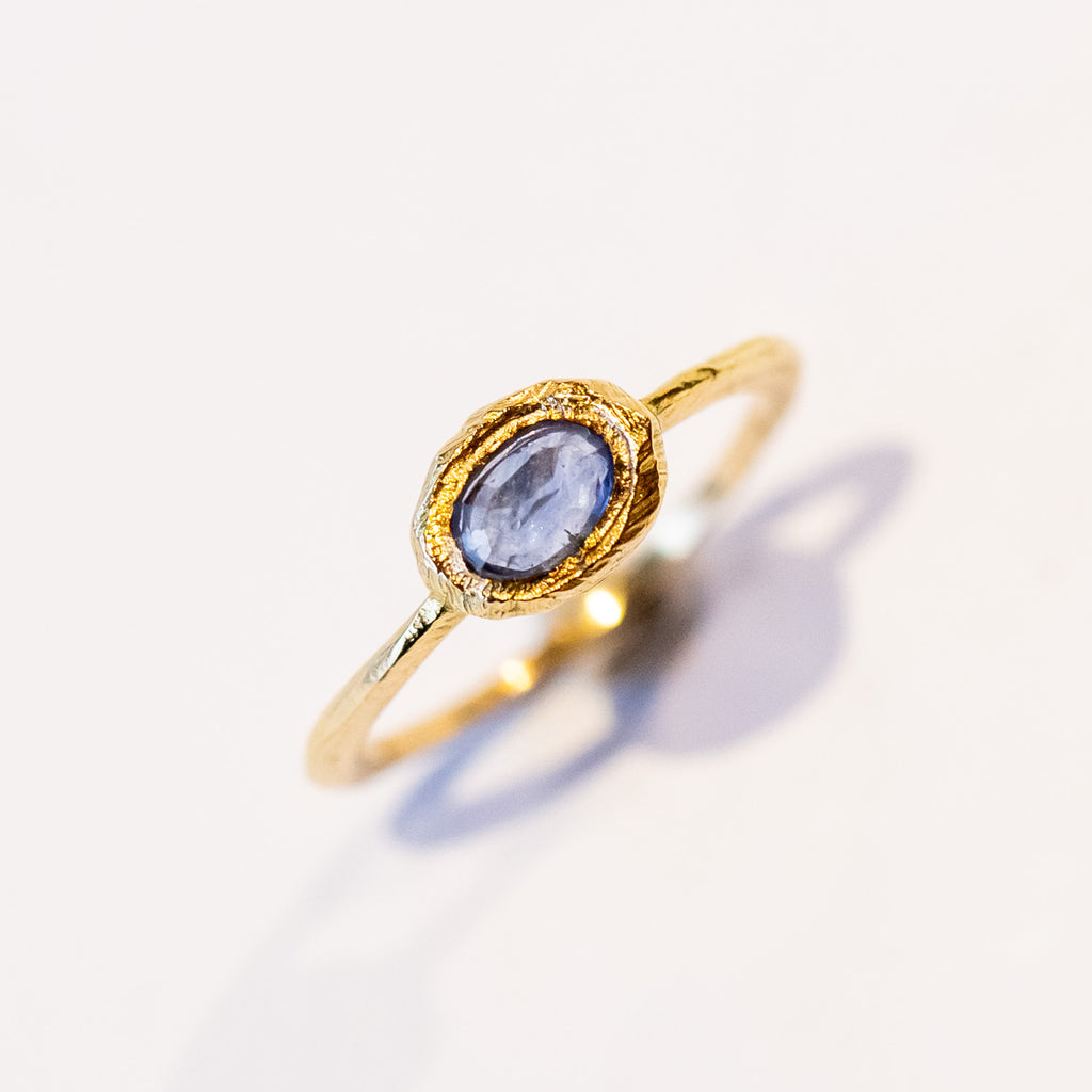 An east-west set oval rose cut blue sapphire is held in a textured yellow gold bezel on a slender ring.