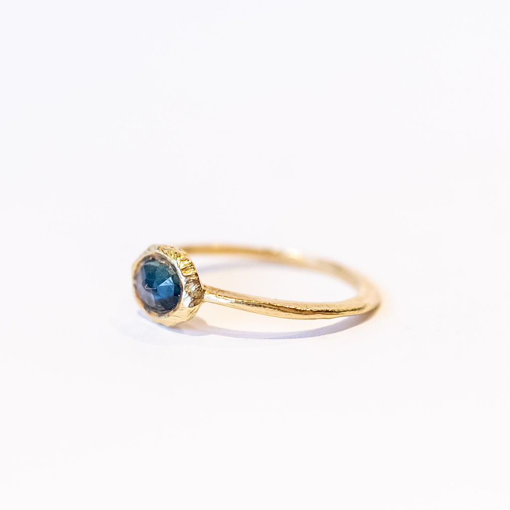 A slender, textured yellow gold ring with an east-west, bezel set teal blue sapphire.