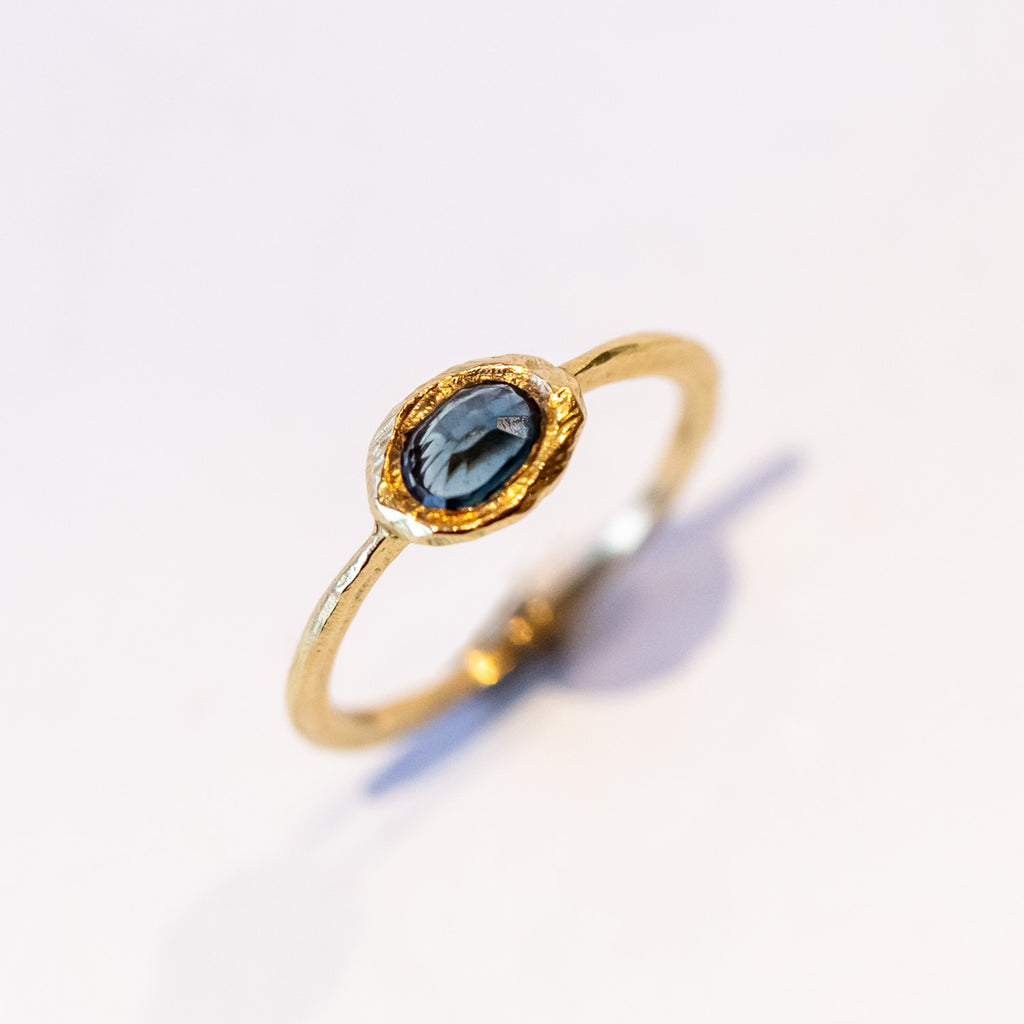 A slender, textured yellow gold ring with an east-west, bezel set teal blue sapphire.