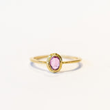 A oval rose cut pink sapphire is set into a textured bezel on a slender yellow gold ring.