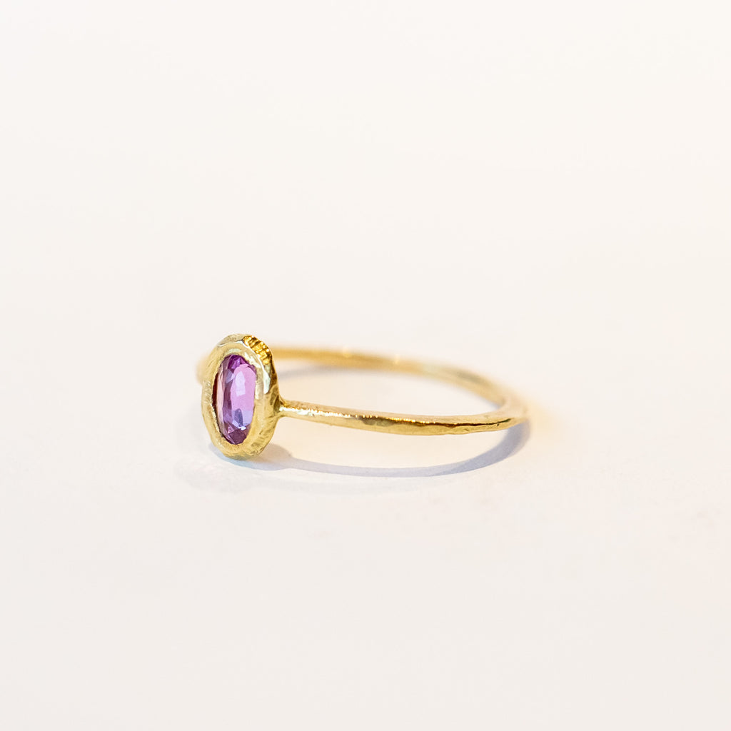A oval rose cut pink sapphire is set into a textured bezel on a slender yellow gold ring.