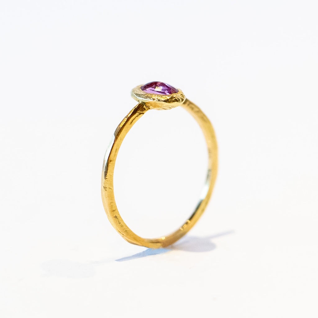 A oval rose cut pink sapphire is set into a textured bezel on a slender yellow gold ring.