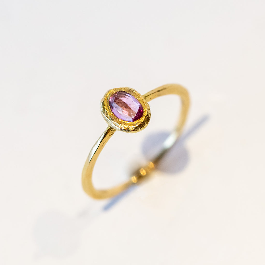 A oval rose cut pink sapphire is set into a textured bezel on a slender yellow gold ring.