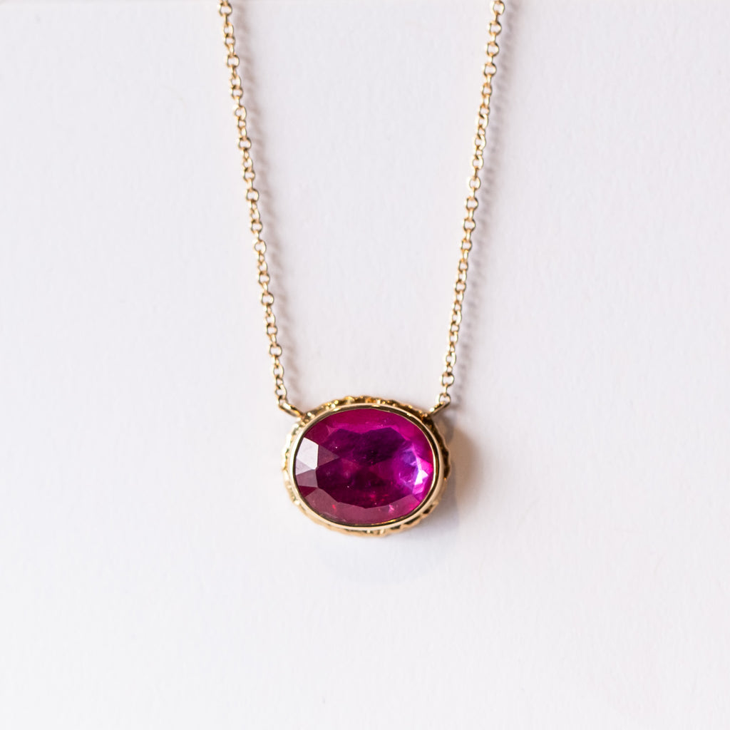 A bright pink, faceted oval ruby gemstone is bezel set in yellow gold stationed on a yellow gold cable chain necklace.