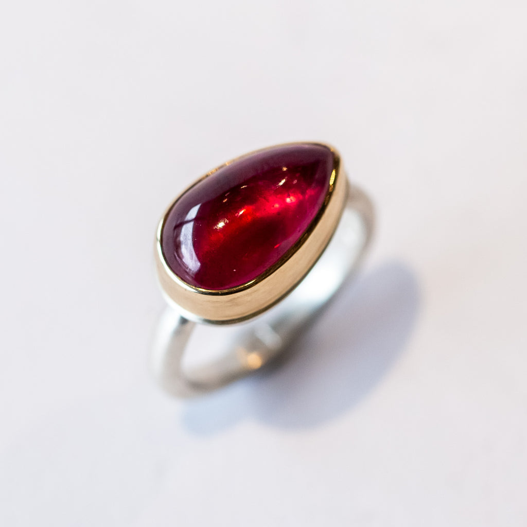An east-west set teardrop shaped African ruby cabochon is bezel set in yellow gold on a silver ring.