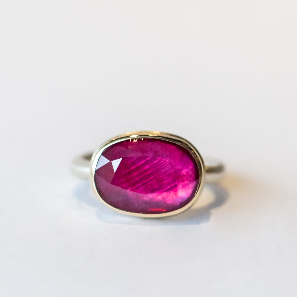 An oval faceted ruby gemstone is set horizontally in a yellow gold bezel on a silver ring.