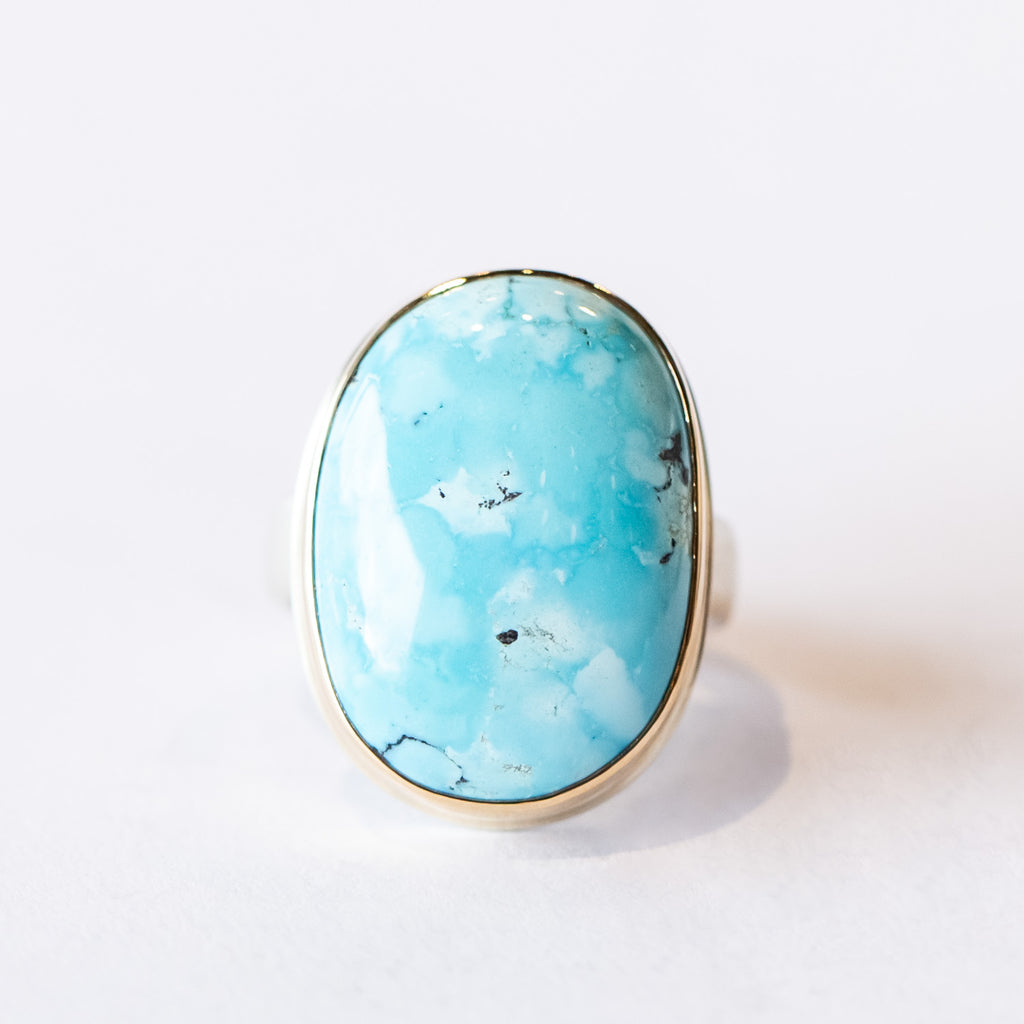 A large oval cabochon turquoise gemstone is bezel set in yellow gold on a silver ring.