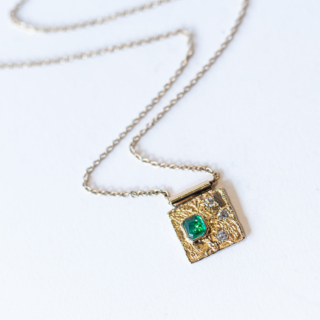 A square cut emerald and three tiny diamonds are set in a square gold textured plaque on a cable chain necklace.