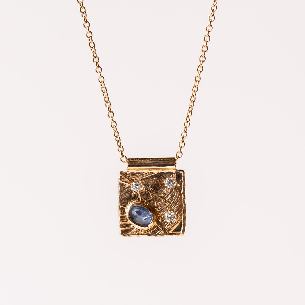 A blue oval sapphire and three tiny diamonds are set into a yellow gold textured square plaque on a cable chain necklace.