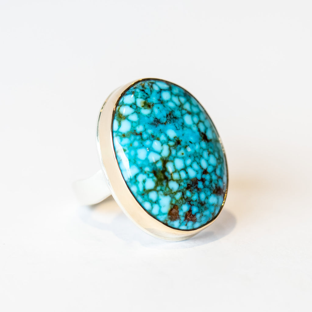A large oval turquoise gemstone is bezel set in yellow gold on a silver ring.