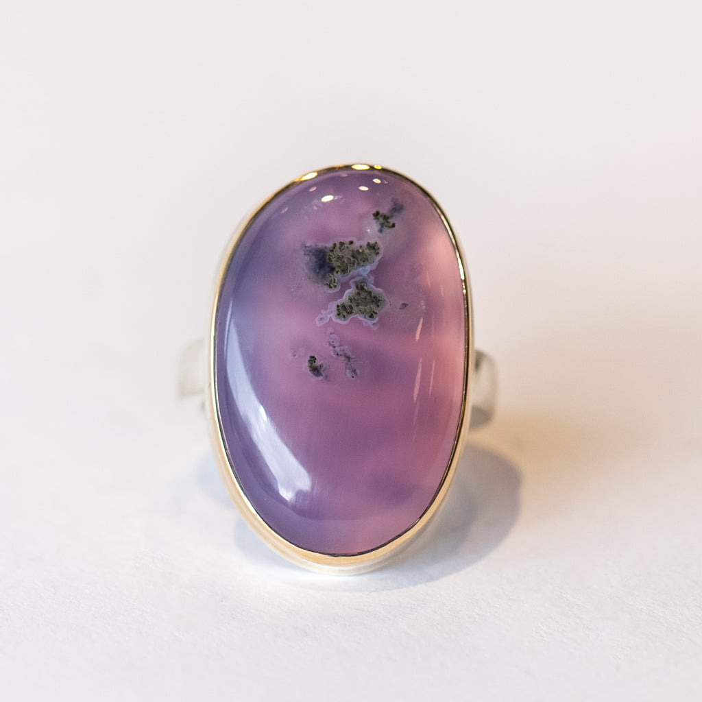 A large oval purple chalcedony gemstone with some visible matrix inclusions is bezel set in yellow gold on a silver ring.