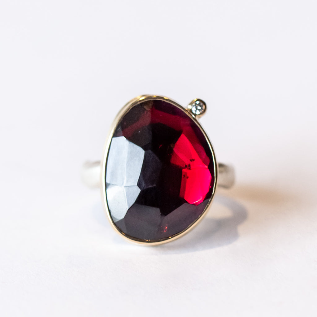 An asymmetrical deep red garnet is bezel set in yellow gold on a silver ring with one tiny diamond accent.