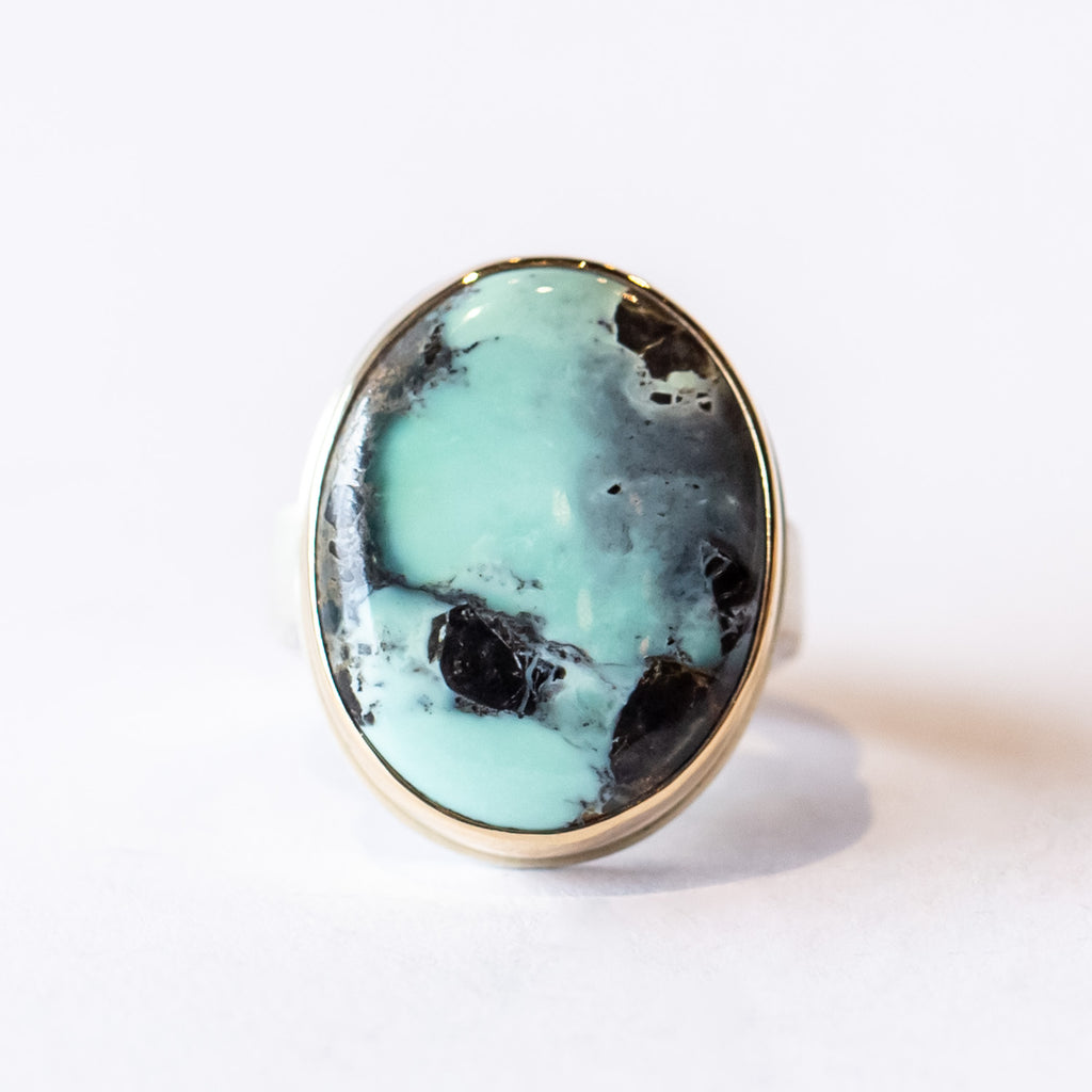 A large oval teal and black turquoise cabochon gemstone bezel set in yellow gold on a silver band.