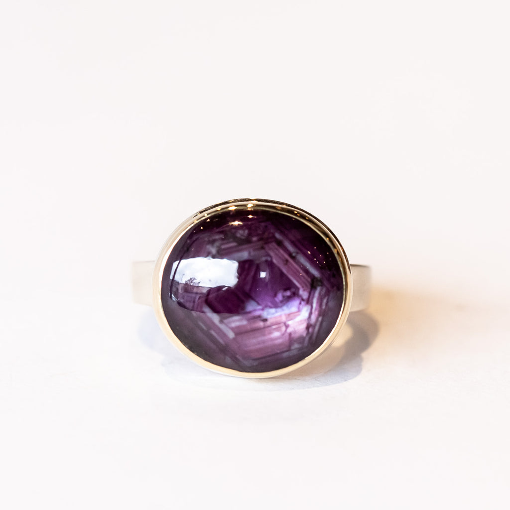 A deep reddish-purple star ruby cabochon gemstone is bezel set in yellow gold on a silver ring.