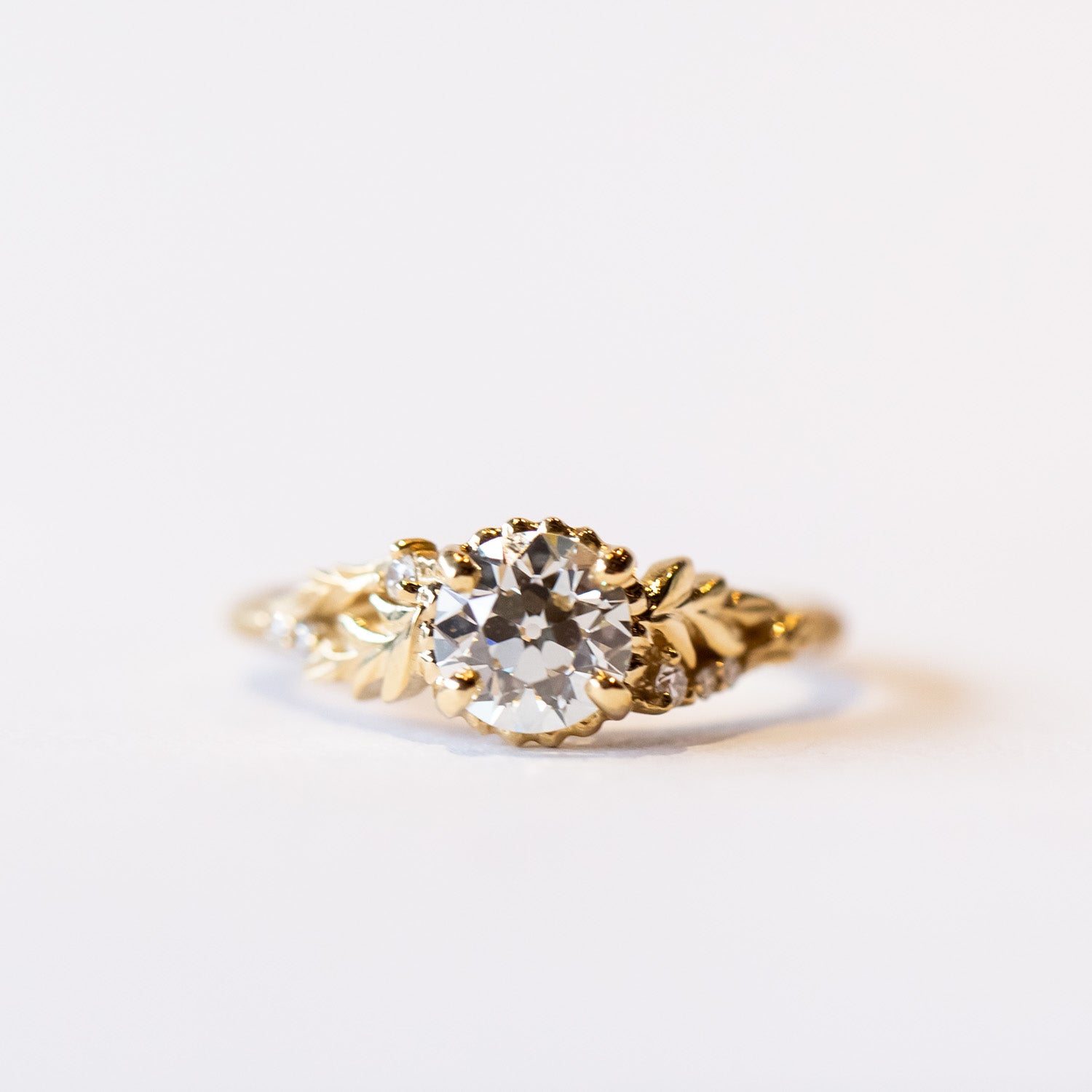 A yellow gold cathedral engagement ring featuring an old European cut center diamond with foliate shoulder accents and tiny side diamonds.