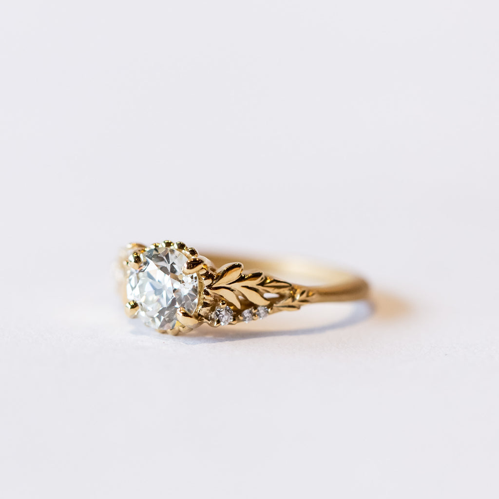 A yellow gold cathedral engagement ring featuring an old European cut center diamond with foliate shoulder accents and tiny side diamonds.