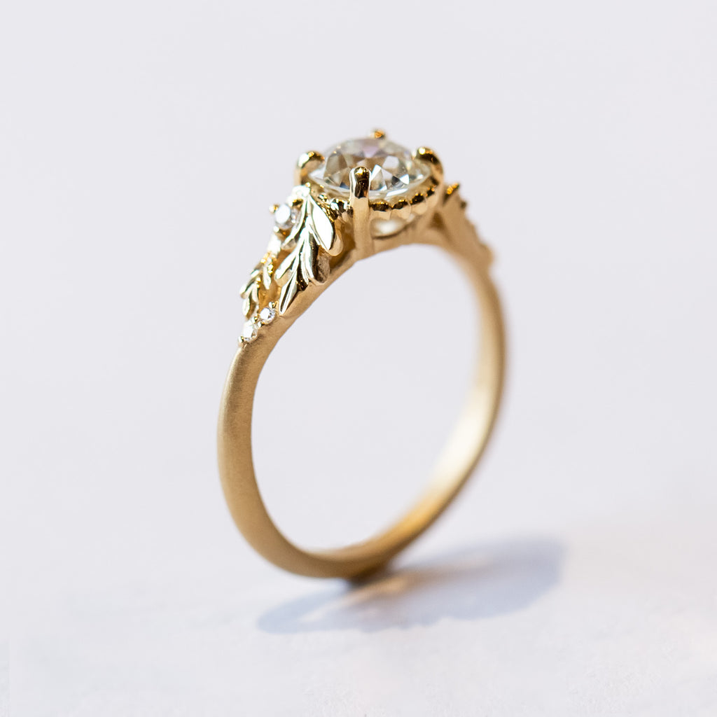 A yellow gold cathedral engagement ring featuring an old European cut center diamond with foliate shoulder accents and tiny side diamonds.