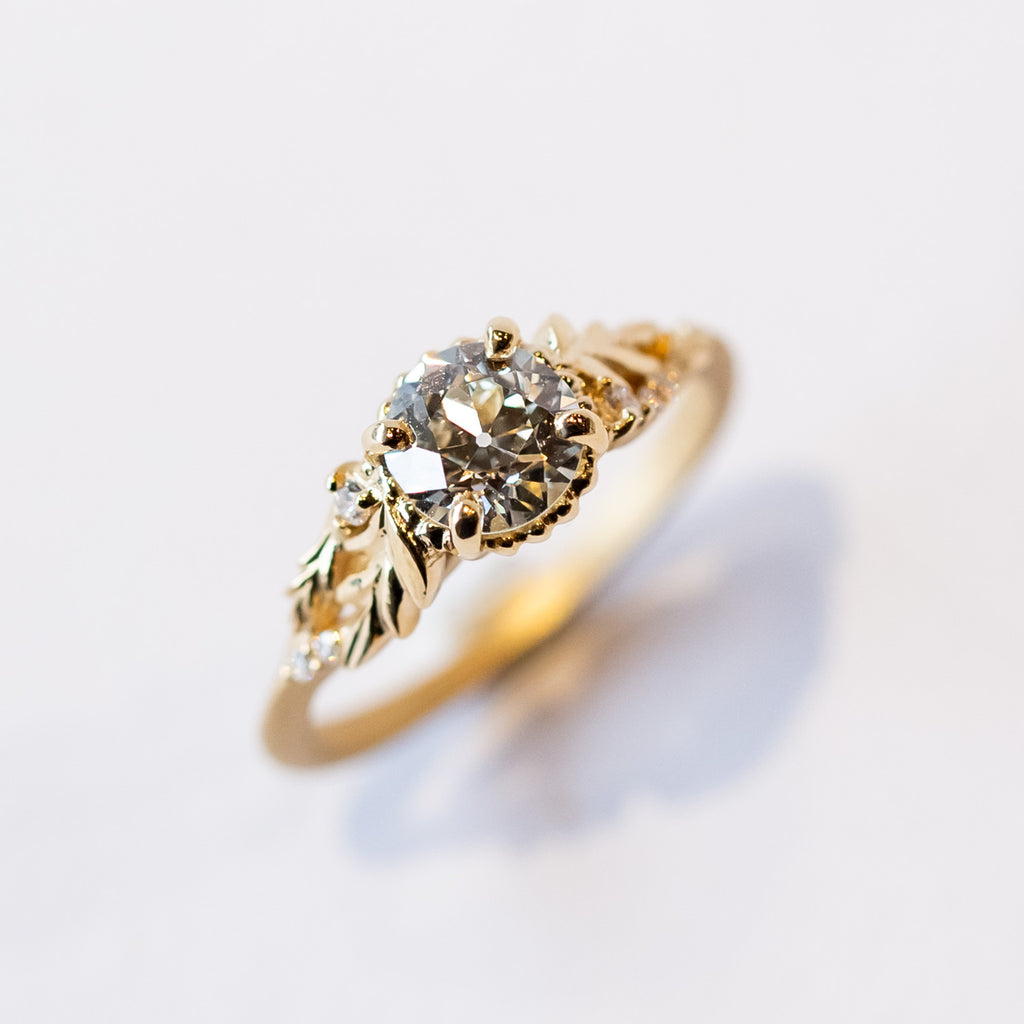 A yellow gold cathedral engagement ring featuring an old European cut center diamond with foliate shoulder accents and tiny side diamonds.