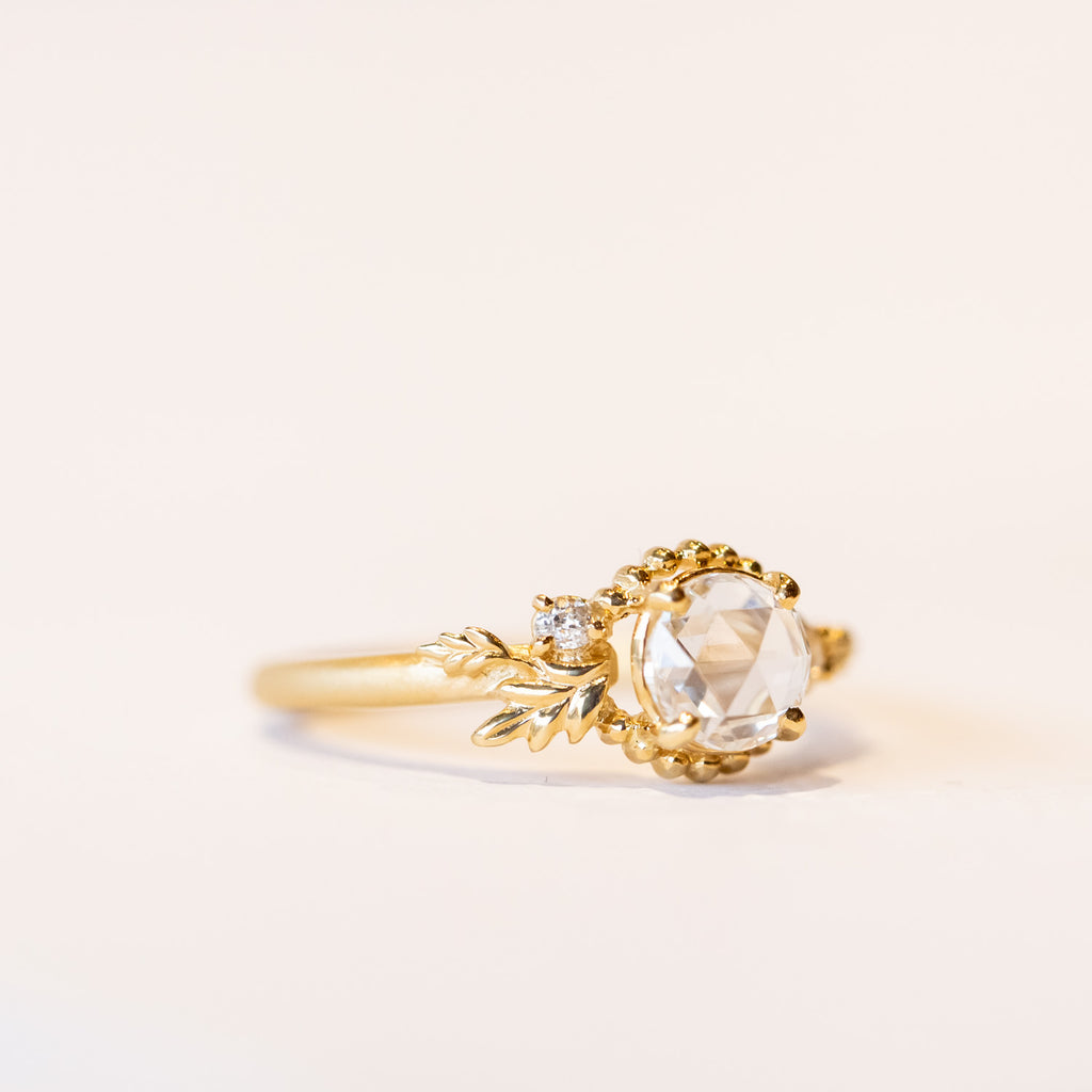 A yellow gold engagement ring with a round, rose cut center diamond accented by asymmetrical foliate elements and two small side diamonds.