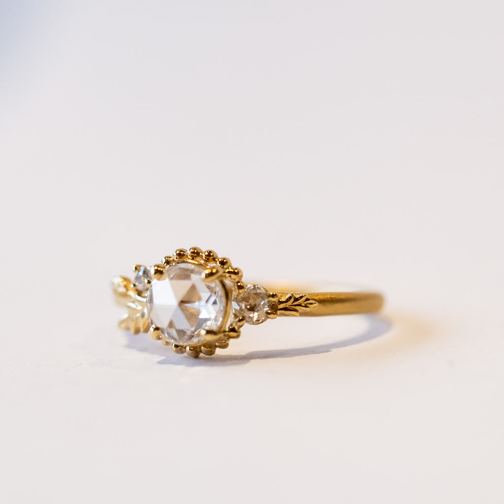 A yellow gold engagement ring with a round, rose cut center diamond accented by asymmetrical foliate elements and two small side diamonds.
