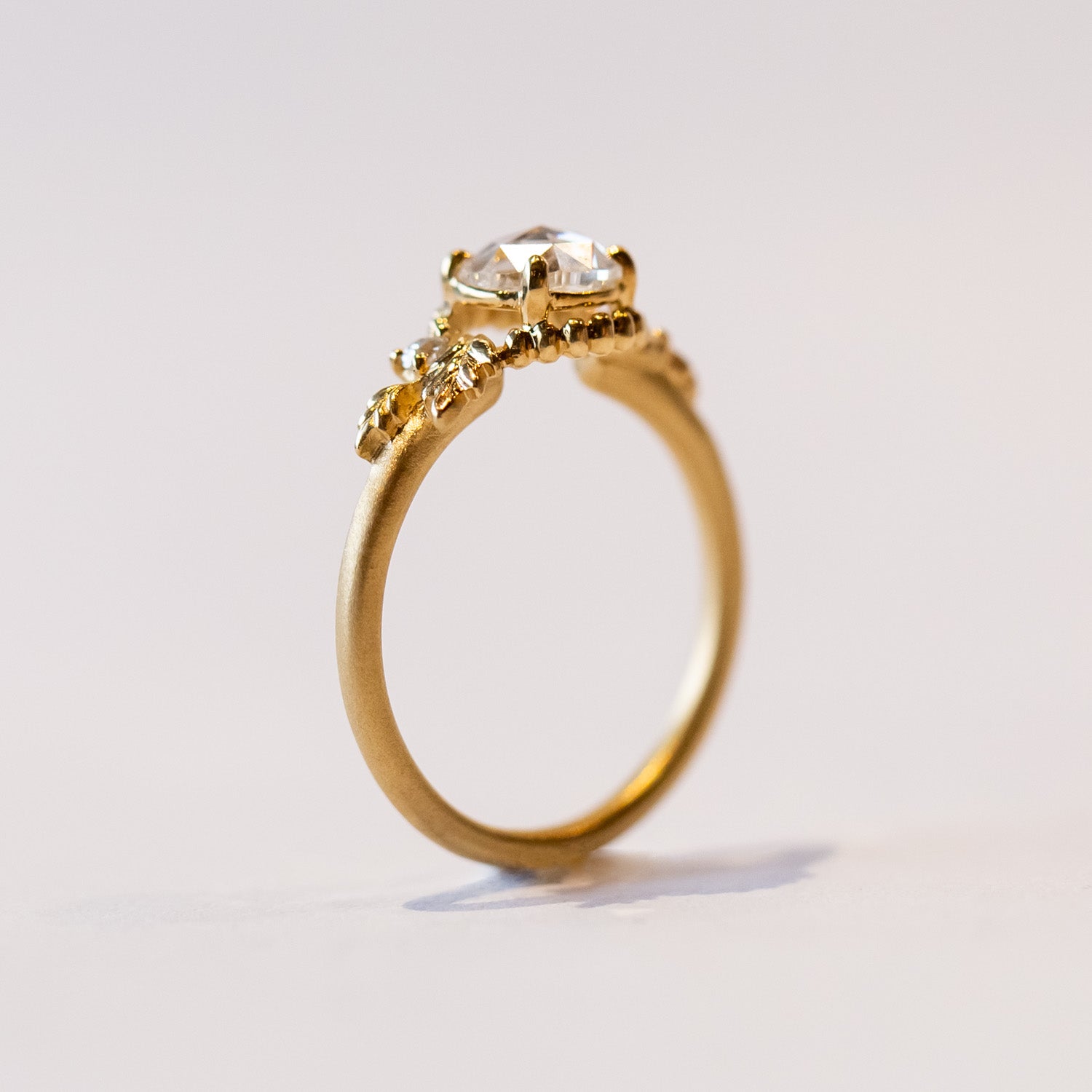 A yellow gold engagement ring with a round, rose cut center diamond accented by asymmetrical foliate elements and two small side diamonds.