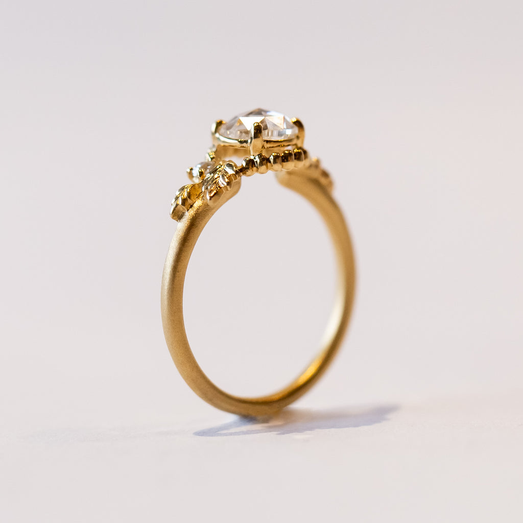 A yellow gold engagement ring with a round, rose cut center diamond accented by asymmetrical foliate elements and two small side diamonds.