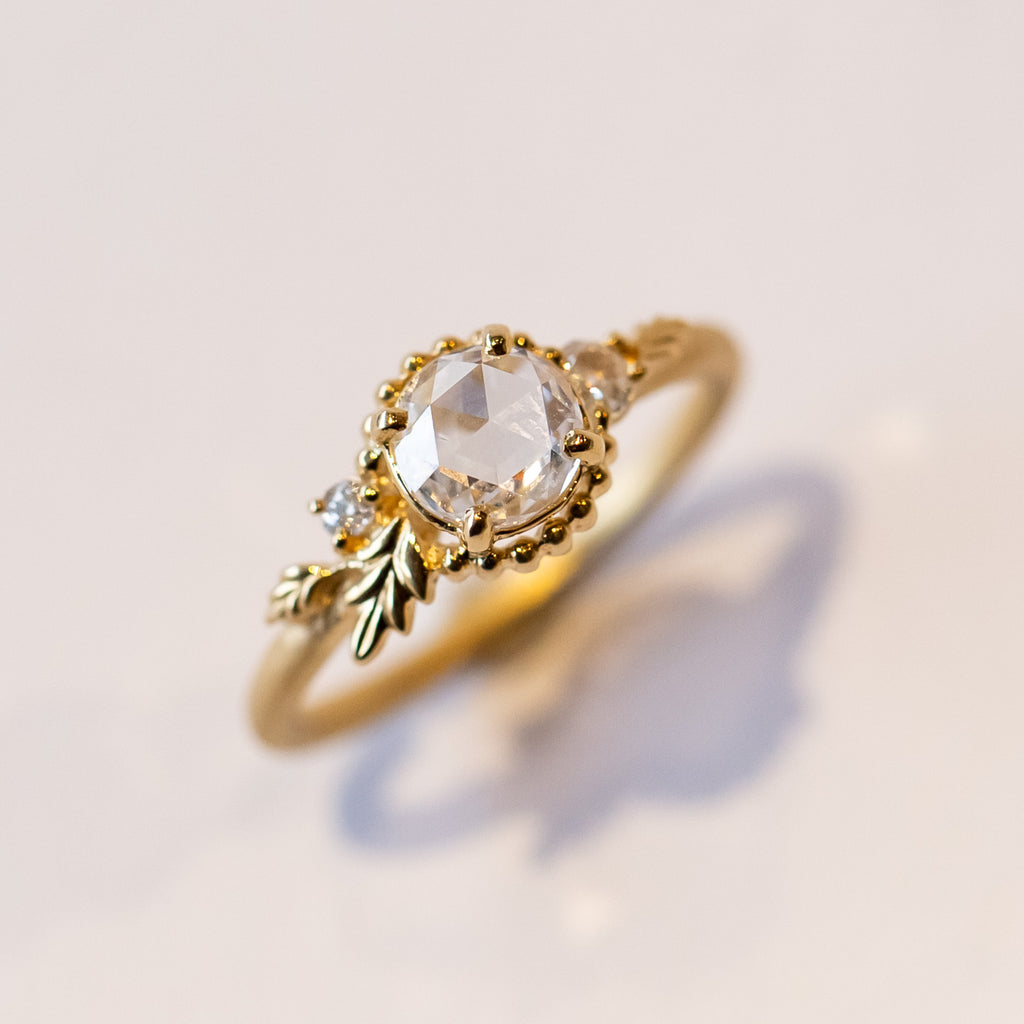 A yellow gold engagement ring with a round, rose cut center diamond accented by asymmetrical foliate elements and two small side diamonds.