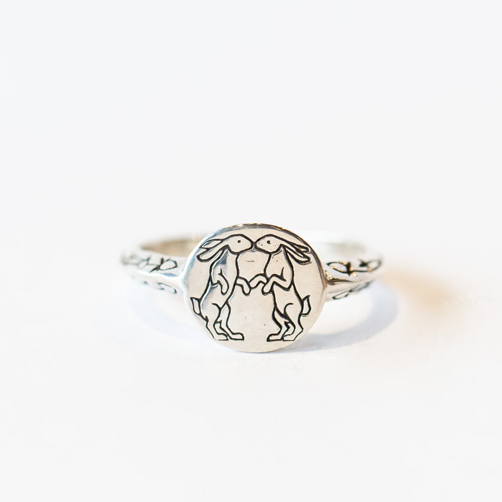 A silver signet ring with two engraved kissing rabbits on a round face and clover along its shoulders.