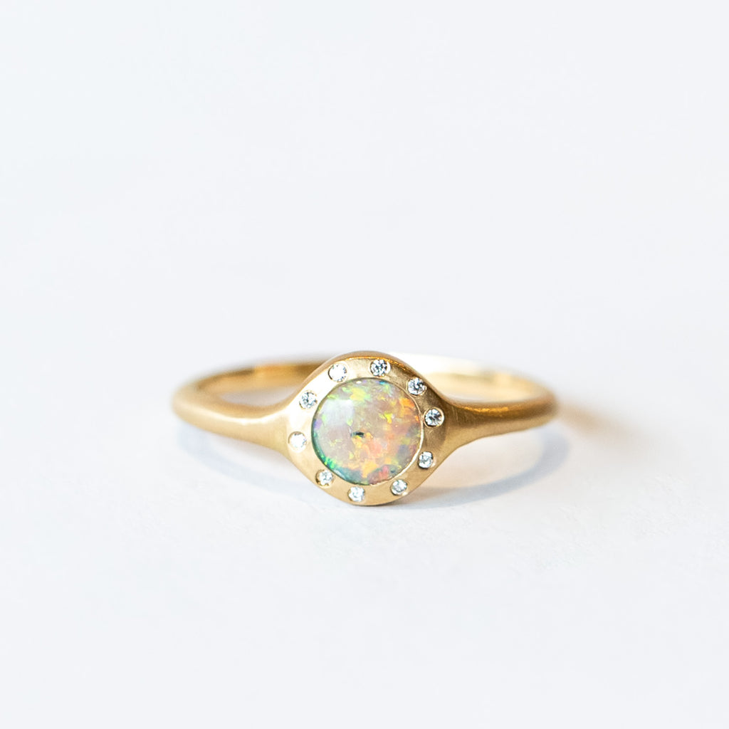 A matte-finished yellow gold dainty signet  ring featuring a round opal flush set in the face, surrounded by flush set white diamonds.